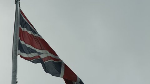 A large Union Jack flag, symbol of Great Britain flutters in a strong wind against a grey sky on a flagpole. 4K