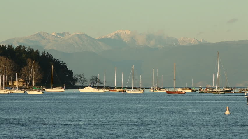 Boats cross Nanaimo Harbor in front of a popular anchorage. A floatplane passes in the sky above. The snow capped Coast Mountains rise in the background. British Columbia, Canada.
