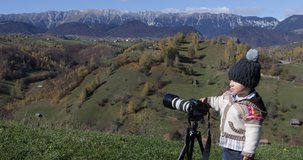 Little kid playing the artist photographer with a photo camera on tripod for shooting gorgeous nature autumn mountain landscape, 4K