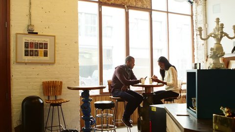Wide shot. A couple sits at a table in a cafe and looks at their phones, not each other