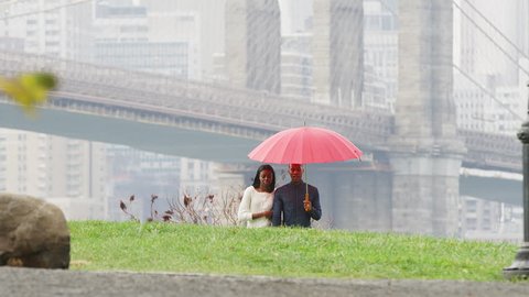 A couple holds hands and an umbrella as they walk through the park towards the camera with the Brooklyn Bridge behind them