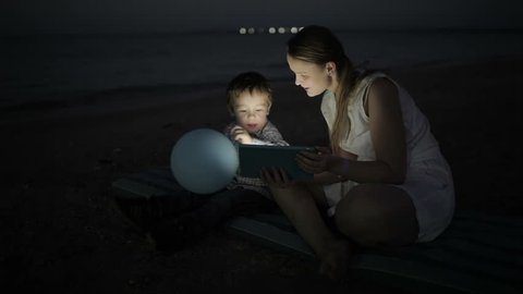Panning shot of a little boy and his mom sitting by the sea with touchpad. Screen enlighting their faces