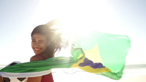 Cheering brazilian woman with flag and football in slow motion at the beach with flare