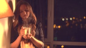 Beautiful Girl Drinking Tea or Coffee at home. Beauty Model Woman enjoying the Cup of Hot Beverage and looking out the window on the night city. Full HD 1080p video footage