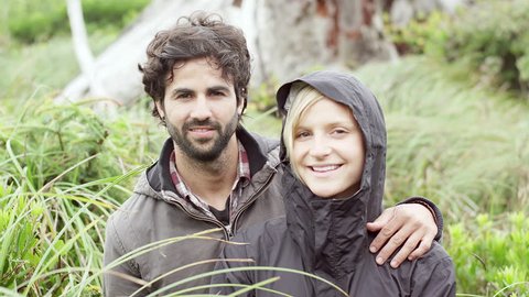 A couple smiles at the camera while in green wooded nature. The man hugs the woman who is wearing a jacket with her hood pulled up.