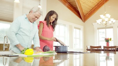 A man and woman stand in the kitchen preparing food. He cuts veggies and she stirs a pot with a spoon
