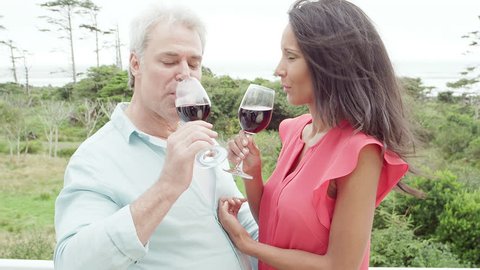 A man and woman stand on the deck and drink wine as they look out to the water and get blown by the breeze