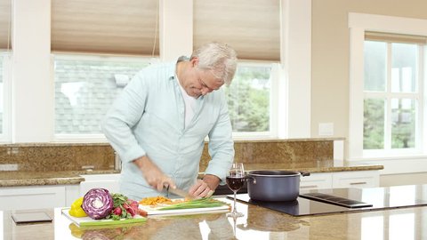 Wide shot. An older man is cutting and preparing food when he gets a phone and answers it while drinking wine