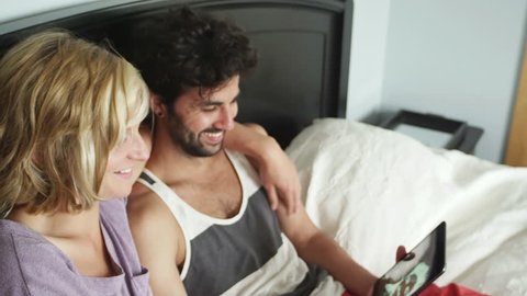 A young couple laying in bed and playing with a tablet look up and smile into the camera