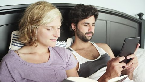 Close up shot of a young couple laying in bed while the woman reads a book and the man looks through his tablet