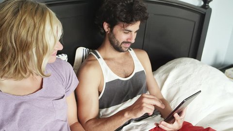 A young couple lays in bed while the man plays with his tablet