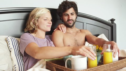 A topless man sits in bed with a woman and they drink juice together