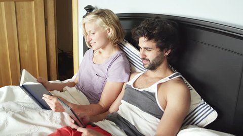A young couple lays in bed while the woman reads a book and the man looks through his tablet when suddenly and female head appears and then floats away