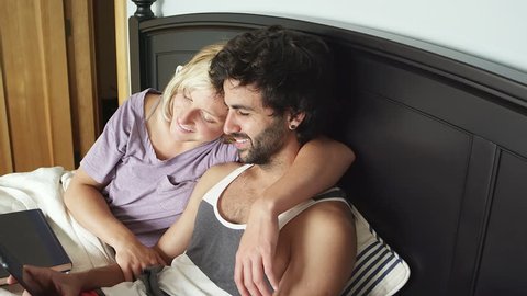 A young couple lays down together in bed and looks through a tablet