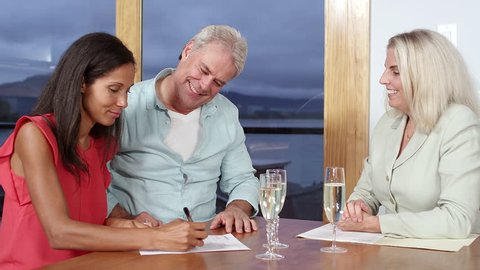 A couple signs some paper work while sitting at a table with a real estate agent and then lift their glasses and toast