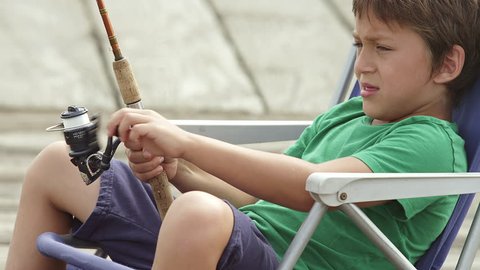 A boy sits in a chair and reels in his fishing line while on a dock