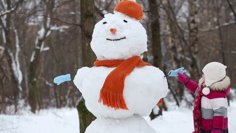 Little girl walk around snowman in orange hat and scarf at winter park
