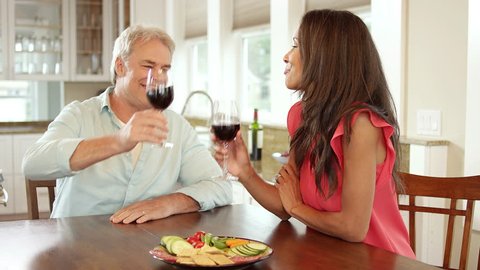 Wide shot of a man and woman walking to the dining room table with a plate of food and drinking a glass of wine