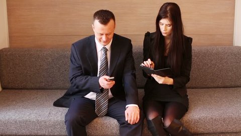 Man and woman in business suits sit on couch with communication device