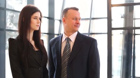 Man and woman in business suits look right and turn faces near big windows