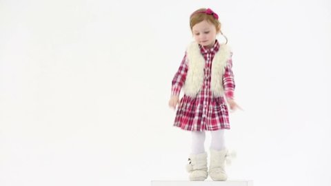 Little girl in fur white vest and boots dances on big white cube