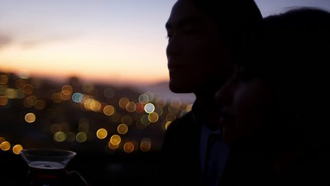 Close up of a man and woman standing on a roof drinking a cocktail at sunset with the city lights behind them