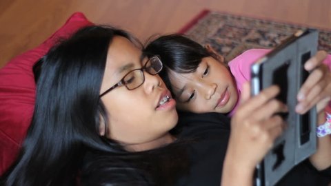 Two cute Asian sisters enjoy spending time together playing on their new digital tablet in the living room.