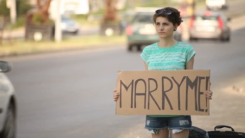 Hitchhiking young adult woman hitchhiker holding Marry Me proposal written board pointing thumb up on interstate highway.