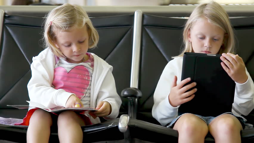 Two young girls sit in chairs waiting, while playing on a tablet and coloring in a book