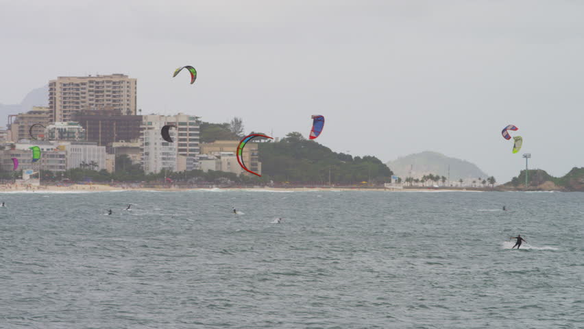 Panning shot from shore of parasailing surfers and cityscape.