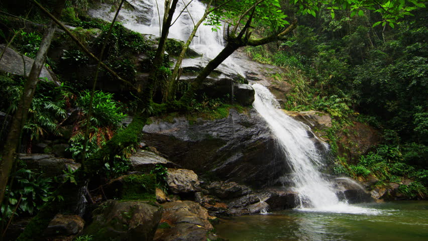 Tracking shot of a jungle waterfall rushing down a rocky hillside into a deep