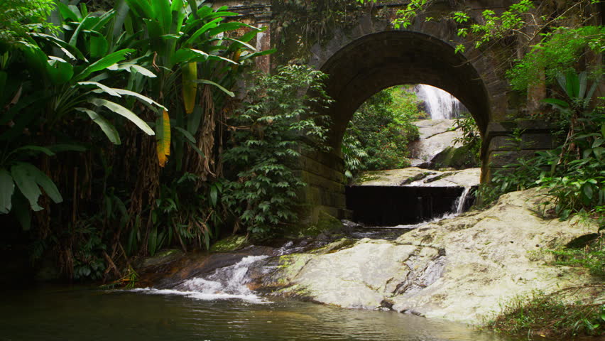 Tracking shot of a scenic stream flowing underneath an arched bridge.