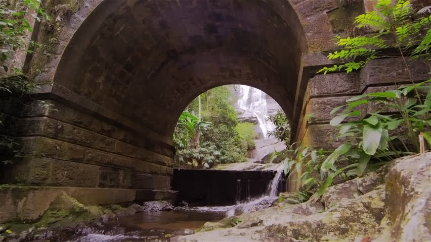 Tracking shot of a stone archway in a jungle with a waterfall beyond.