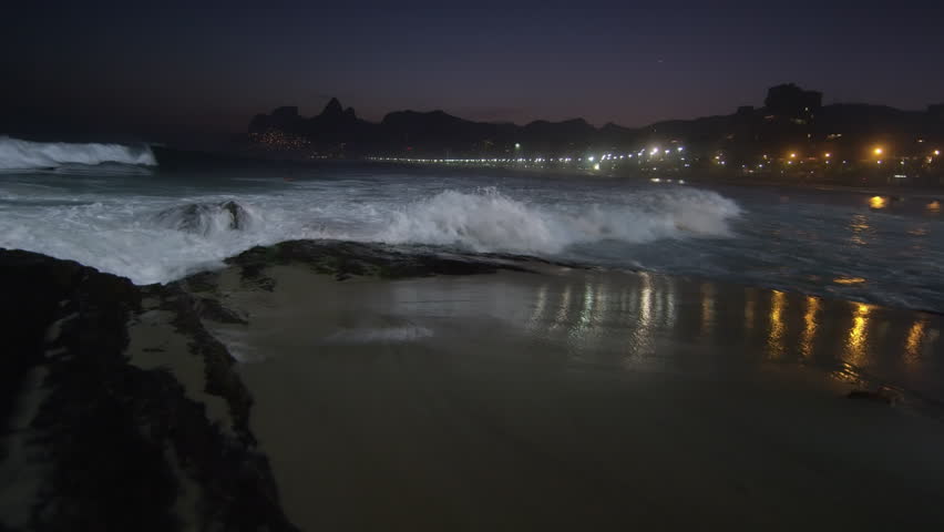 Slow motion, pan right as the tide flows up on Ipanema beach at night