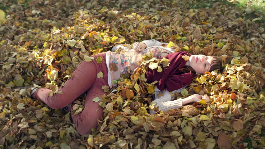 A young woman laying in a pile of dead leaves catches them as they fall from the sky while laughing, in slow motion