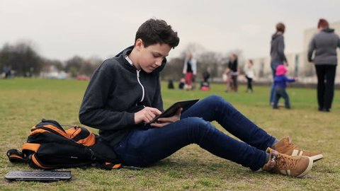 Young teenager with tablet computer sitting on grass in park
