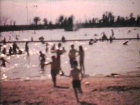 A shot of a crowded beach with tons of kids playing in the water and then a little boy runs towards the camera. (Vintage 8mm film)