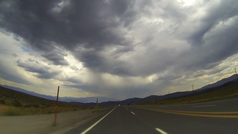 Roadtrip Rearview Time-lapse. The camera looks back on the winding road trailing behind the vehicle as storm clouds billow overhead in the outskirts of rural Nevada.