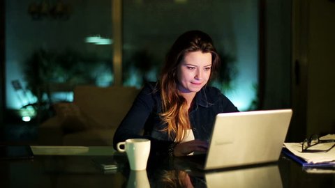 Woman working on laptop at night at home and drinking coffee.

