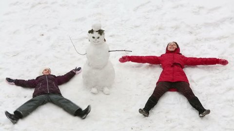 Mother with daughter lie on the snow near snowman and shake hands and legs like a butterfly