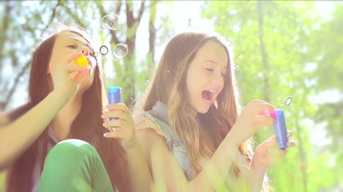 Beauty teen girls having fun outdoors. Beautiful joyful teenagers laughing and blowing soap bubbles in spring park. Girlfriends outdoor. Slow motion video footage 1080 full hd. High speed camera shot