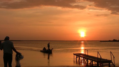 Video of fishing boats and water skier in distance at Chock Canyon Lake in south Texas.  Vivid bright orange sunset. Fisherman throwing a net for bait fish.