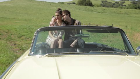 Front view of a young couple sitting in the back of a vintage convertible car looking at nature and talking to each other