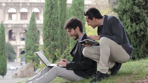 Two business people with tablet and laptop talk about a project on a park
