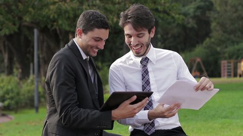 Two young businessmen with tablet computer talk about a project on a park
