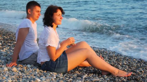 young woman and man sitting in pebble beach near sea 