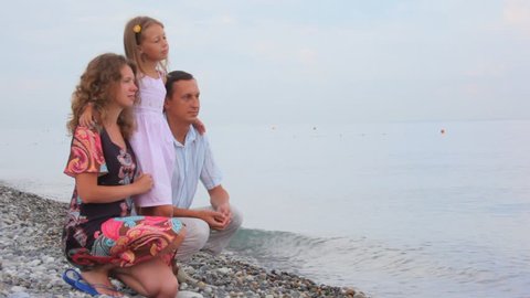family sitting on pebble coast and looks on sea 