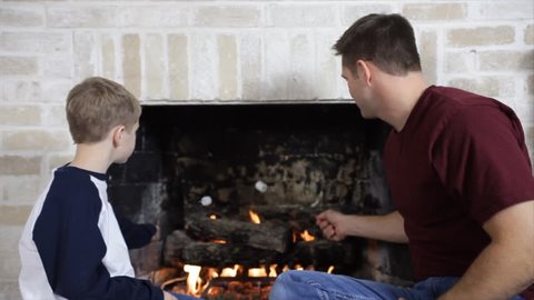A father and his young son, sitting next to the fireplace, marshmallows.