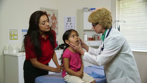 A Caucasian physician checks the ears of a cute little Hispanic girl while her mother looks on with concerned interest.