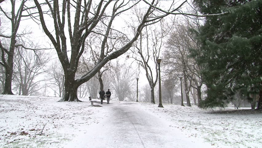 Two people walk city park path during snow fall in Portland, Oregon. Rack focus.